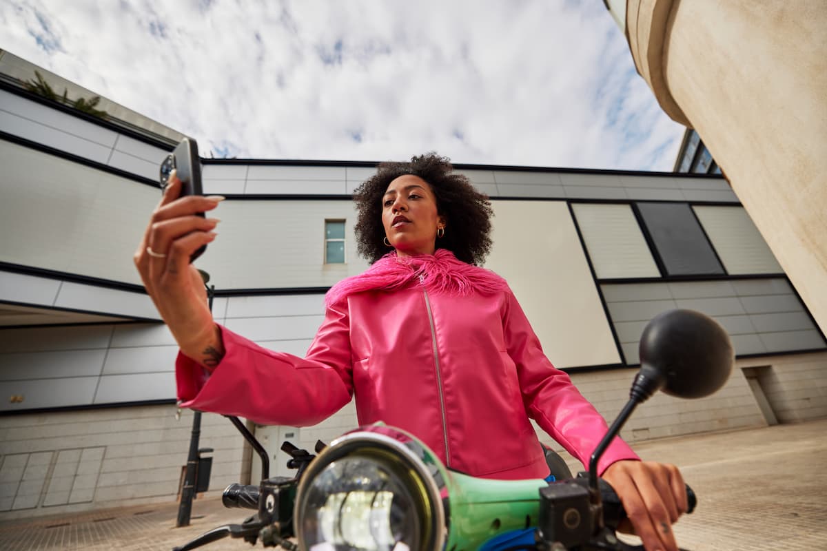 Low angle of young African American female looking at screen of mobile phone while sitting on scooter holding handlebar with hand and taking selfie against buildings and cloudy sky