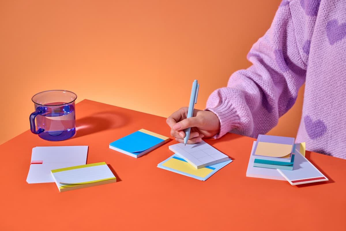 A person's hand writing on a small notepad with a pen, along with a glass of water and other stationary on the desk.