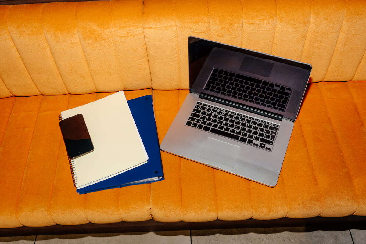 Laptop with pen, smartphone, and notepads placed on cushioned orange in modern interior